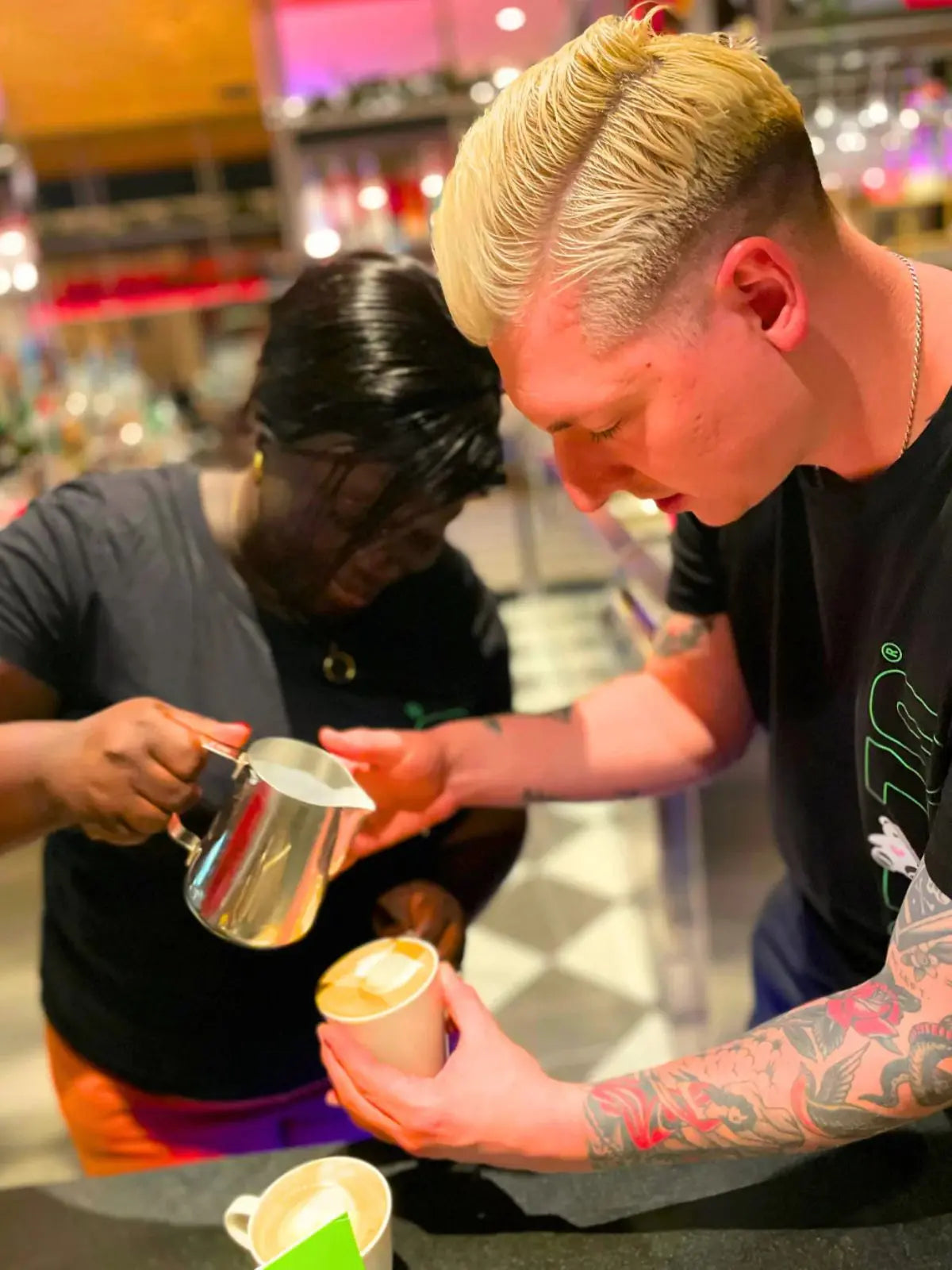 Mozzo US coffee training: Woman holding milk jug pouring into a takeaway cup held by a blonde tattooed man